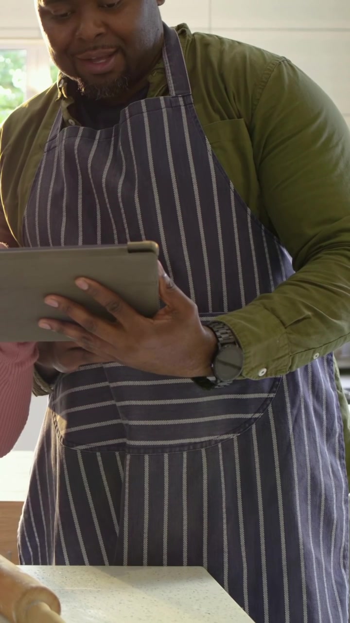Vertical video: Reading recipe on tablet, man in striped apron reaching for rolling pin in kitchen