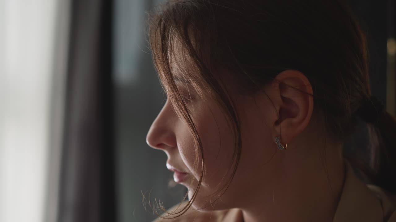 Jovem mulher triste pisca o olho olhando pela janela da sala de estar.