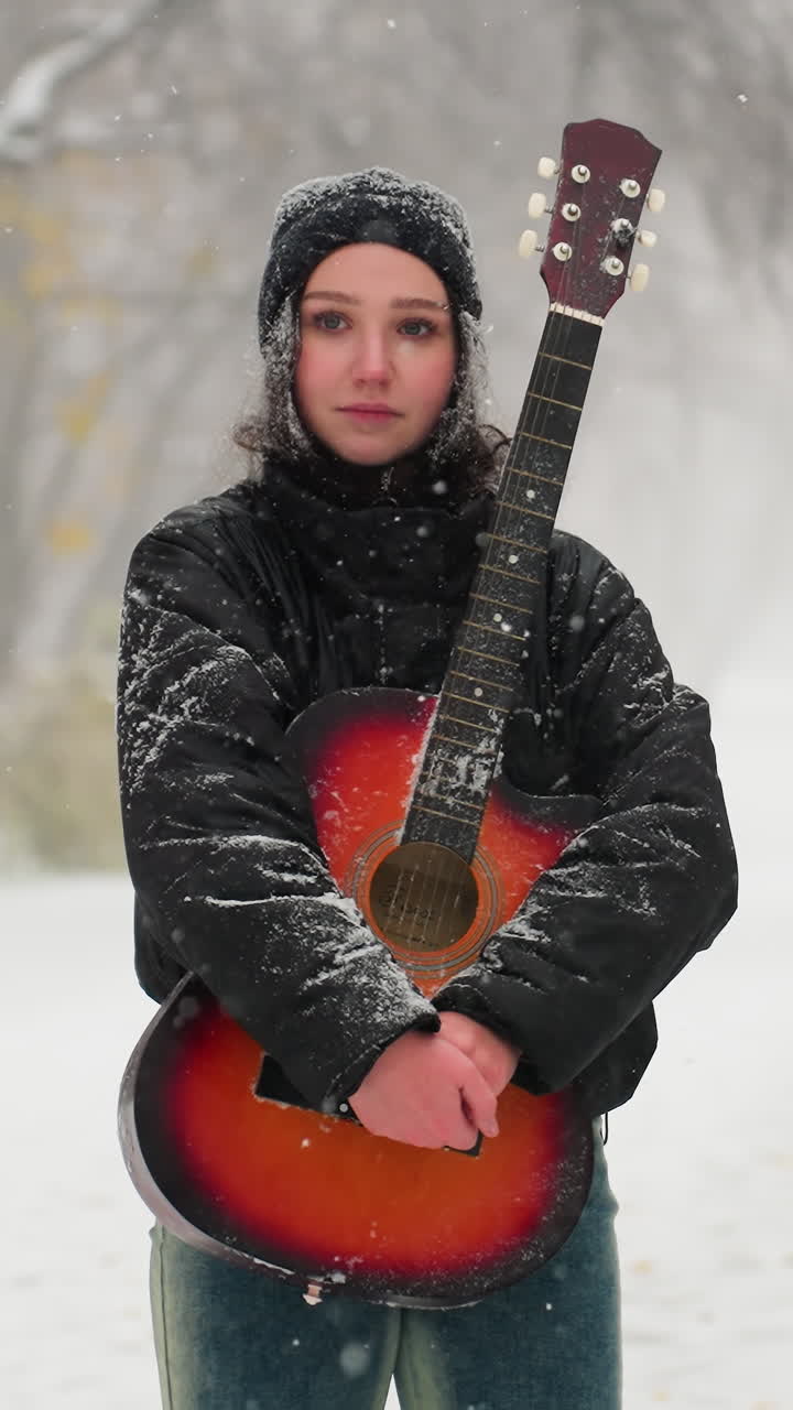 joven de pie en un parque nevado sosteniendo una guitarra acústica roja vibrante, rodeada de árboles helados y nieve cayendo suavemente y vista borrosa de personas en la lejanía