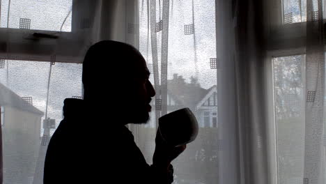 standing beside the window, a man savors his coffee while gazing at the daytime scene outside, exemplifying the essence of routine household moments