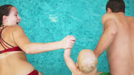 Back-view-of-young-mother-and-father-together-with-their-child-jumping-in-the-swimming-pool-holding-hands.-Mother-and-father-are-teaching-their-kid-how-to-swim