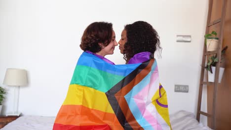 Happy-diverse-young-friends-with-LGBT-flag-touching-nose-tips-sitting-in-bed