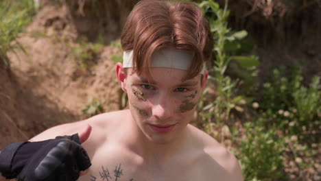 bare chest adventurer in bandage headband with muddy camouflage stripes, turning gently amidst green brown foliage, subtle smile, thumbs cutting throat gesture conveying silent warning