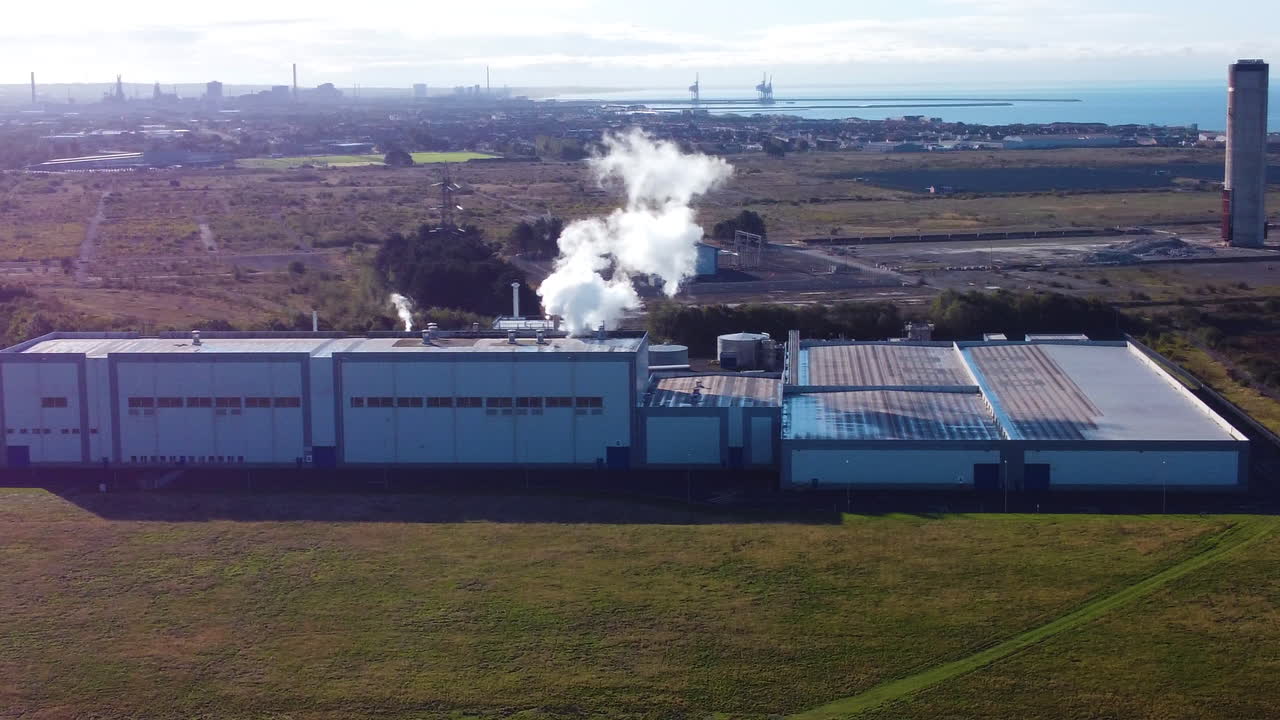 Sideways Aerial View of Factory with Smoke Emissions Rising from Funnel Over Industrial Landscape with Sea Port in Background. ProRes 4K