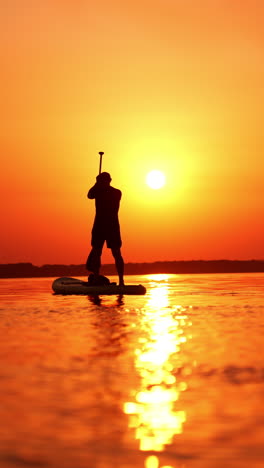 Sportive male silhouette on the orange sunset backdrop. Man practicing sup boarding on the river in summer. Vertical video