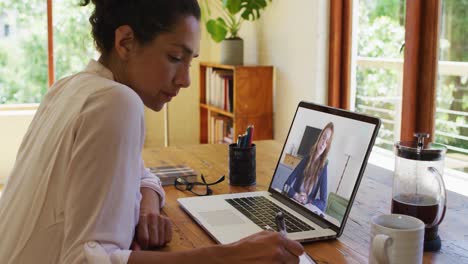 mujer afroamericana tomando notas mientras tiene una videollamada en una computadora portátil en casa