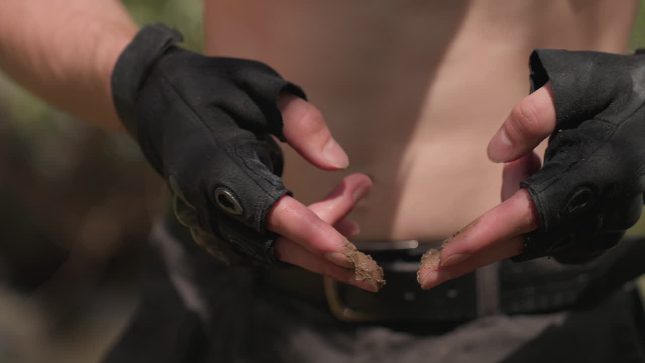 Close-up of shirtless young man wearing tactical gloves while rubbing wet sand between fingers outdoors, showcasing muscular arms and chest tattoo in sunlight near forested riverbank setting
