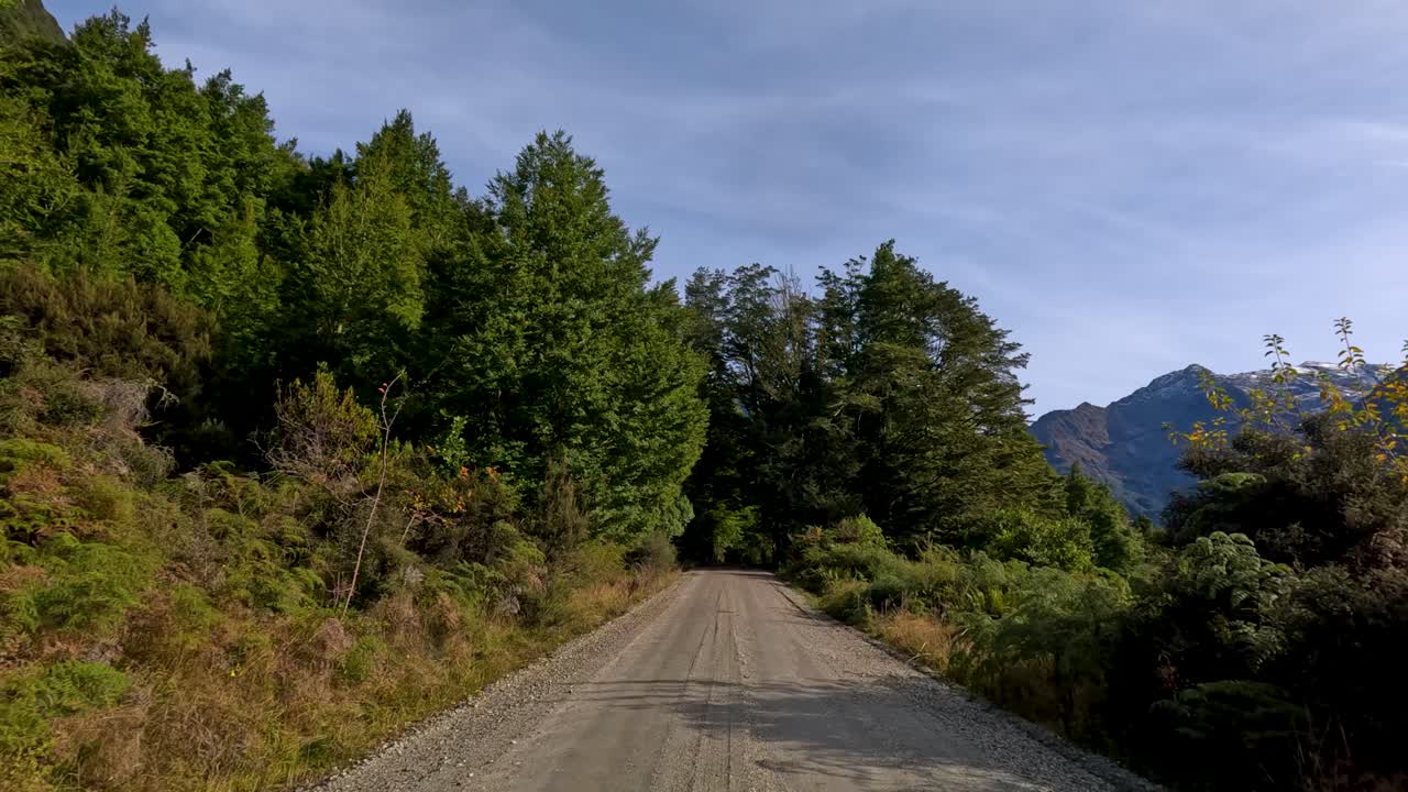 Vehicle travels along gravel road through lush forest, mountain views, natural daylight, steady forward motion
