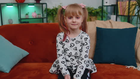 Cheerful-lovely-caucasian-little-toddler-kid-girl-smiling,-looking-at-camera-sitting-on-sofa-at-home