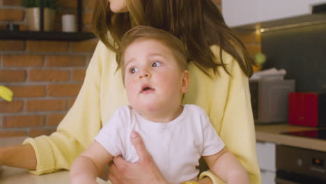 close up view of a baby held by his mother on her lap in the kitchen while she is working on the laptop