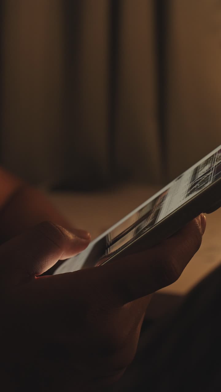 Mano che tiene uno smartphone e scorre un feed di social media in una stanza buia con una scrivania sullo sfondo, illuminata da una luce dai toni caldi. Dipendenza digitale e consumo sconsiderato di contenuti.