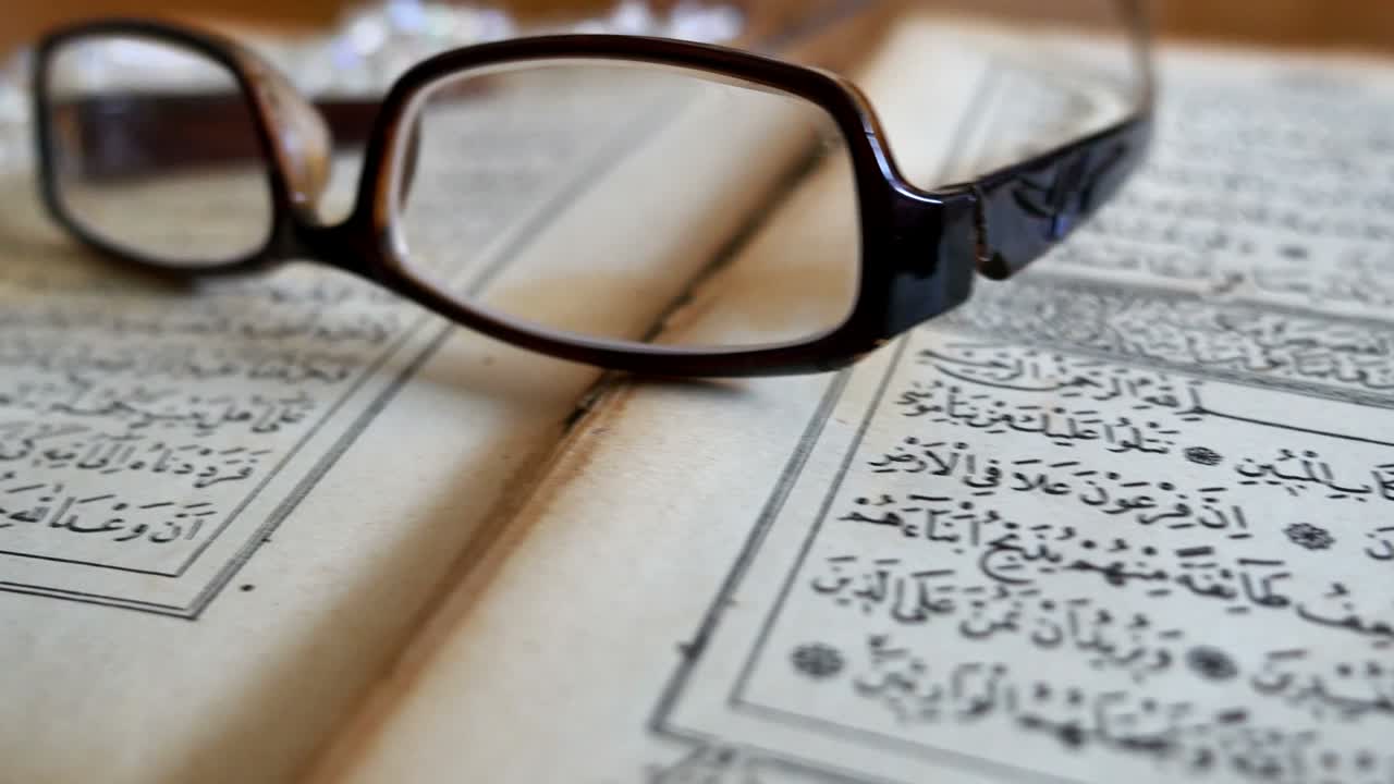 Footage of a Quran lying on a table with reading glasses on it and a tasbeeh in the back, the shot is moving from the right to the left