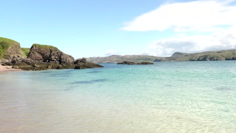 Kristallklare-Wellen-Schlagen-Sanft-Gegen-Den-Sand-An-Einem-Abgelegenen-Schottischen-Strand-Auf-Der-Insel-Handa,-Während-Die-Kamera-Schwenkt,-Um-Grüne-Klippen-Und-Das-Wunderschöne-Türkisfarbene-Wasser-Des-Atlantiks-An-Der-Westküste-Zu-Enthüllen