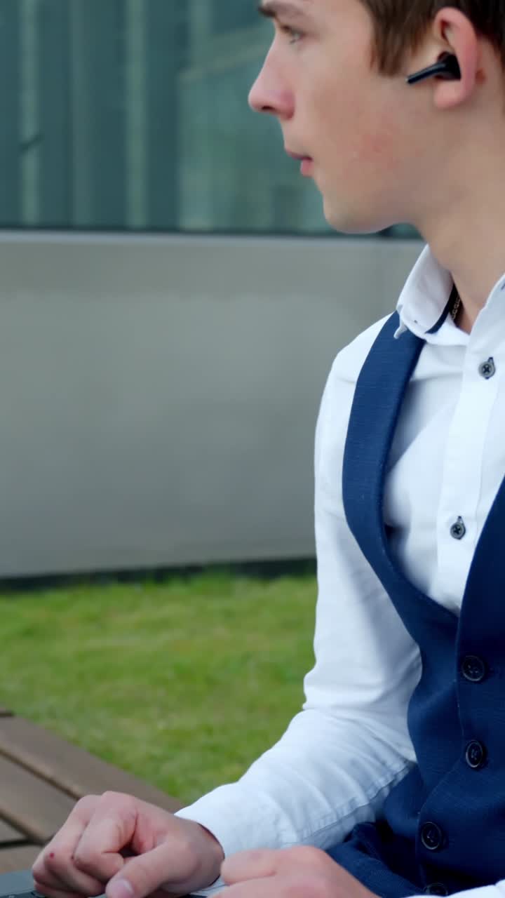 Caucasian young professional man in blue business vest and white shirt, using laptop outdoors while on call wearing Bluetooth earbud, thoughtfully following and agreeing during a business discussion