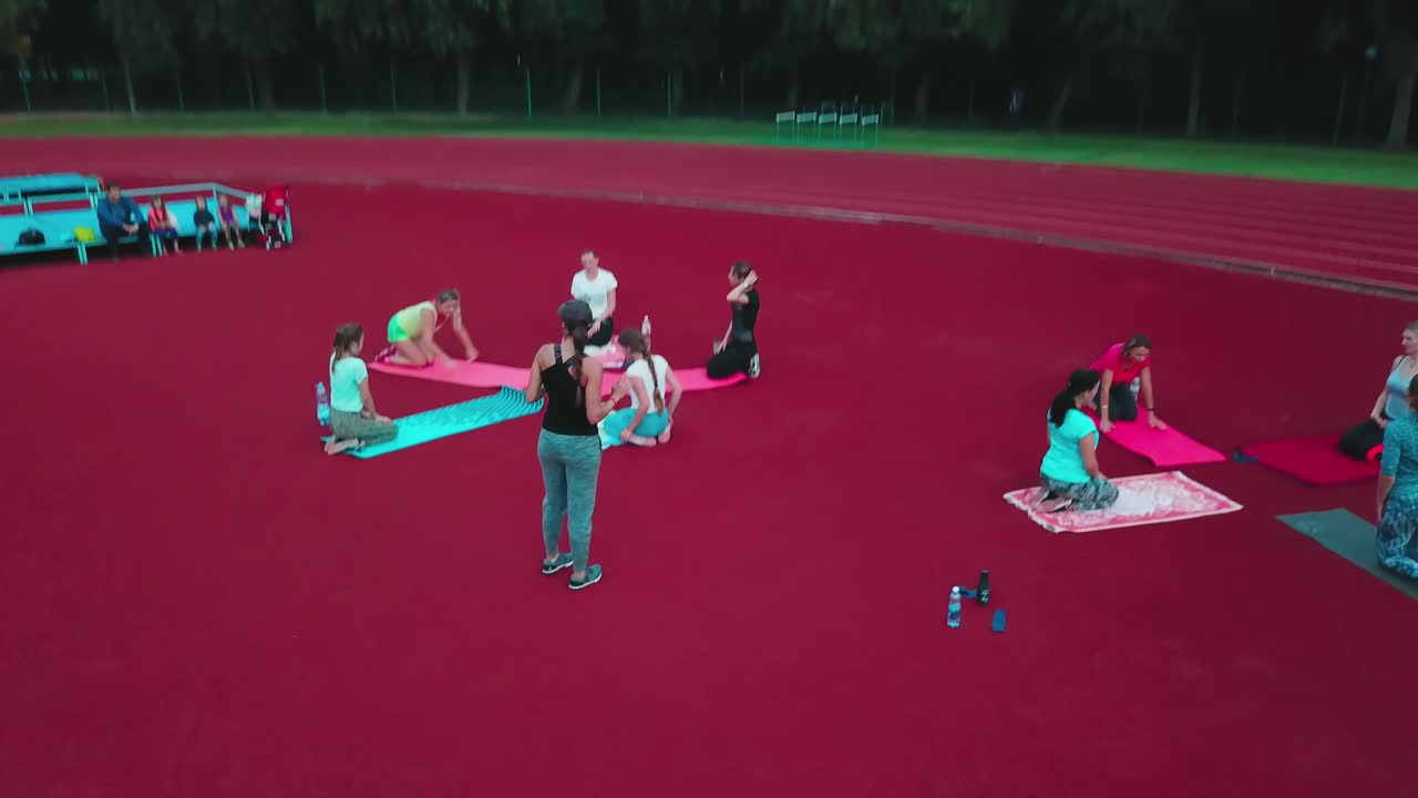 Group of women sitting on mats in the stadium. People doing exercises outdoors in the sports ground.