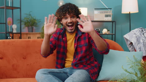 indian man smiling friendly at camera, waving hands gesturing hello, hi, greeting at home on sofa