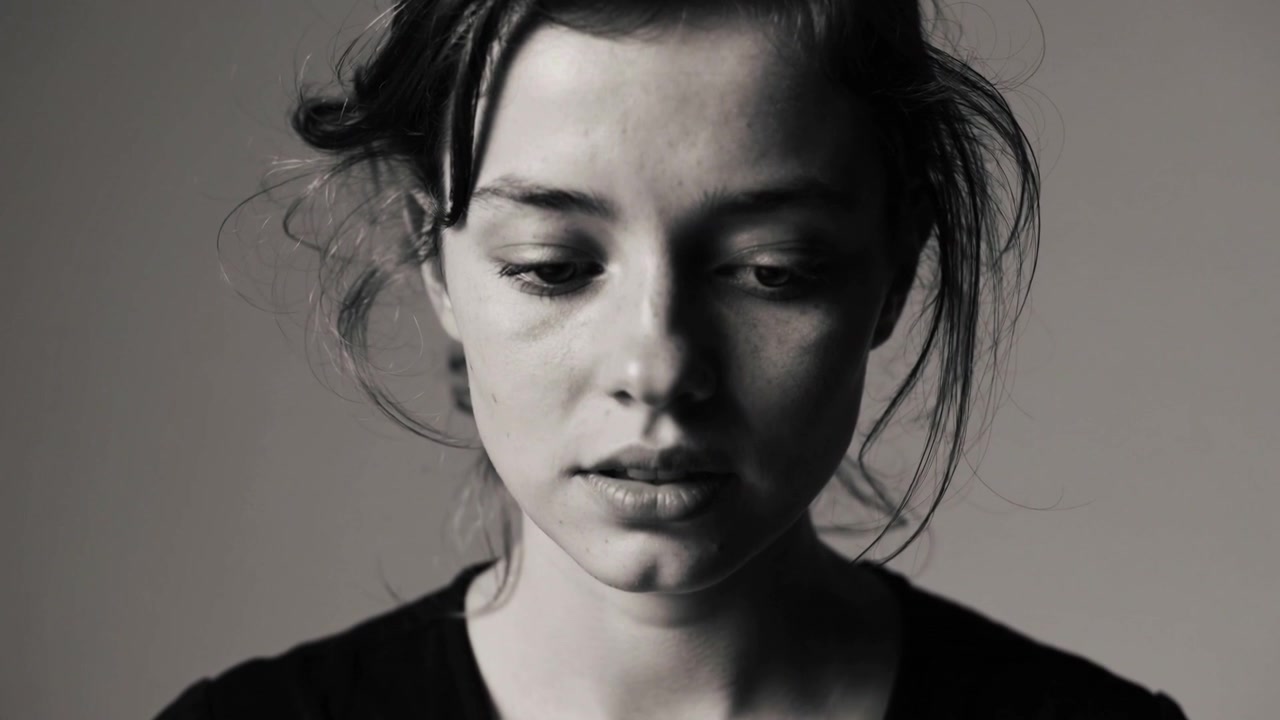 Camera rolling woman holding lowered gaze, parting lips in studio, plain dark top conveying calm