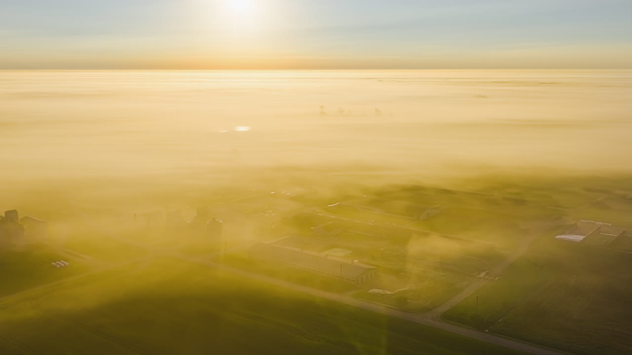 Wispy clouds obscure farmland below dancing and glowing yellow green orange from sunrise