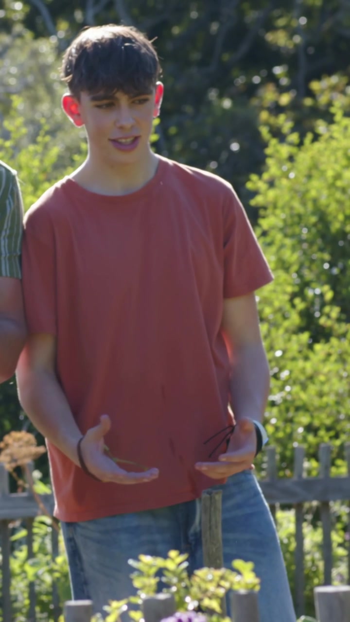 Vertikales Video: Teenager in rostfarbenem Hemd reagiert auf eine Geste, verlagert sein Gewicht und stützt die Hand auf einen Pfosten im Garten.