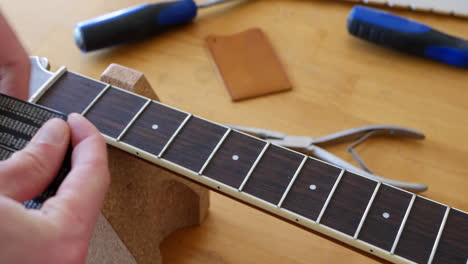 Cerrar-Las-Manos-De-Un-Luthier-Artesano-Midiendo-Y-Nivelando-Un-Diapasón-De-Cuello-De-Guitarra-Acústica-En-Un-Banco-De-Taller-De-Madera-Con-Diapositivas-De-Herramientas-Lutherie