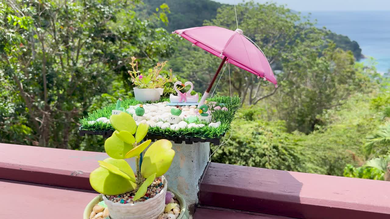 A vibrant garden setting with swan ornaments under a pink umbrella, overlooking lush greenery and ocean views in Phuket