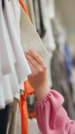 dama de ropa rosa navegando a través de una variedad de ropa colgada en una tienda, ella toca y siente la textura de una camiseta de naranja y ceniza mientras observa otras ropas colgando en el fondo