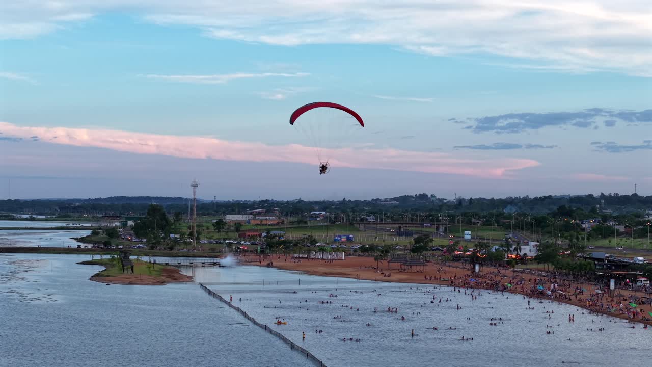 Paralotniarz z napędem lecący nad Balneario Costa Sur w Posadas Misiones Argentyna, ukazujący zatłoczoną plażę rzeczną, szeroką rzekę Paraná, wieczorne niebo, rekreacyjną działalność lotniczą i wypoczynek nad wodą