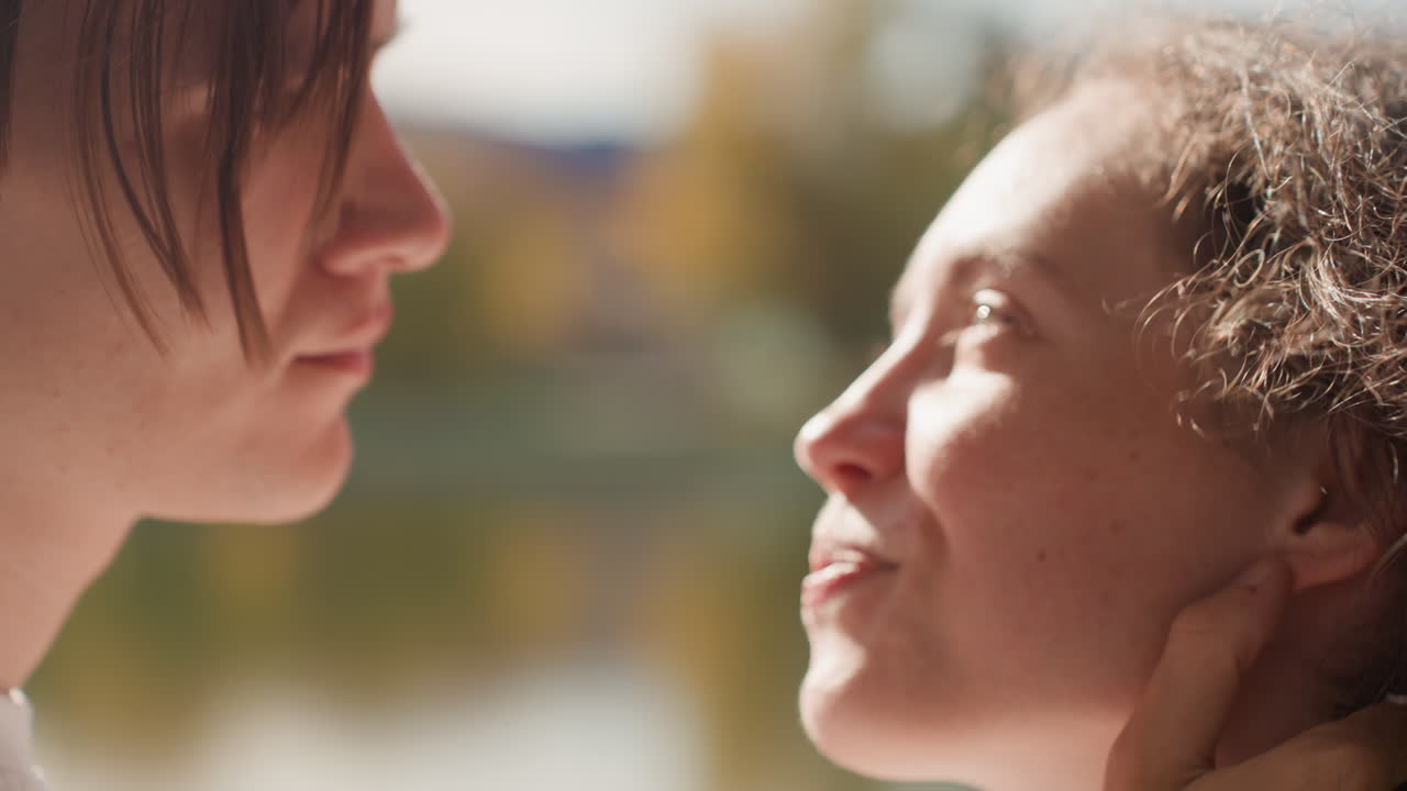 Caucasian couple closeup by lakeside sunlight, young students exchange tender gaze before kiss, hands cradle jaw, soft bokeh reflection on water, intimate engagement mood hinting marriage and future