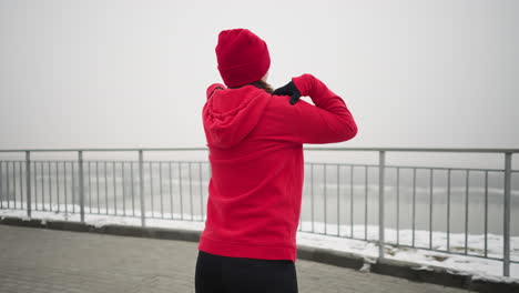 back view of woman in red hoodie and beanie outdoors performing arm stretches by lifting her hands to shoulders and turning her arms background features misty atmosphere with a river and iron railing