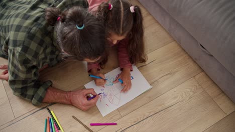 top view of a little brunette girl in a pink dress draws on a white sheet of paper together with her lonely father, a brunette man in a green checkered shirt, using multi-colored markers while lying on the floor near a gray sofa in a modern apartment