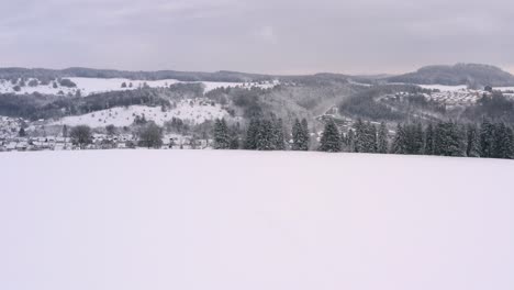 Smooth-flight-over-a-winter-landscape-with-a-wide-white-snow-covered-field-forward-to-the-view-over-a-little-town-behind-a-hill-in-a-dreamy-valley-called-Albstadt-in-Germany