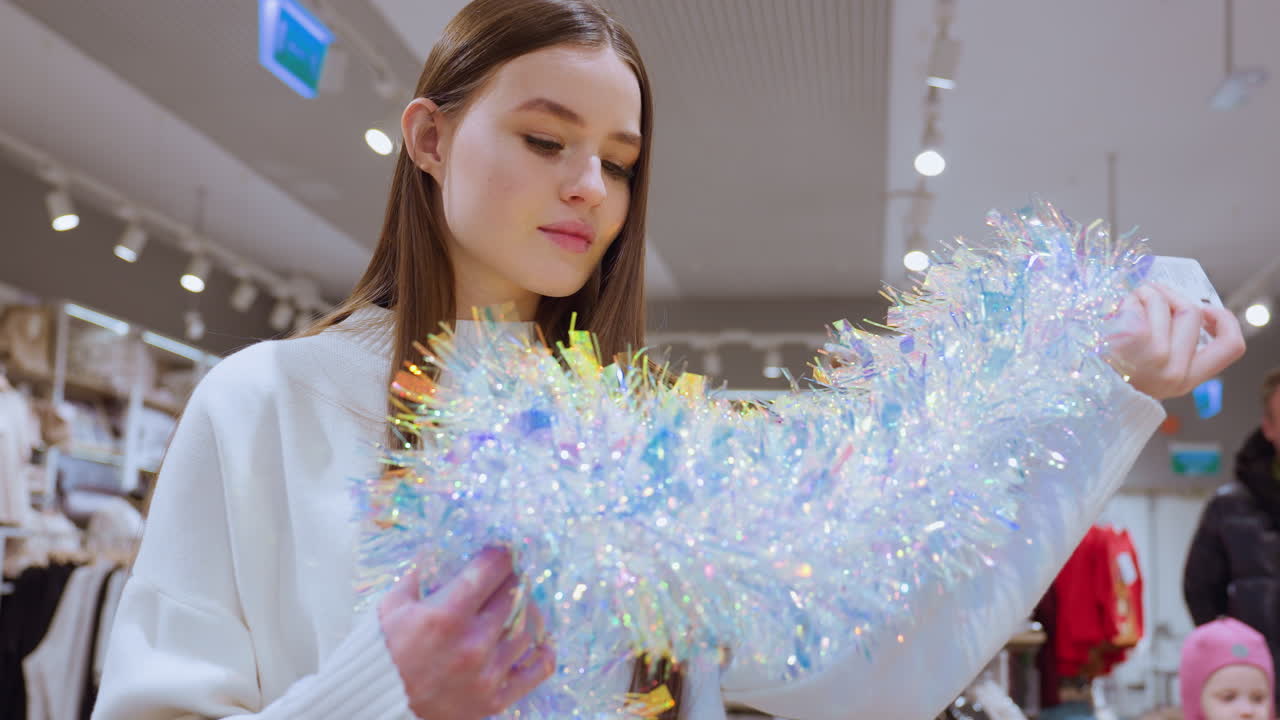 Female shopper holding shimmering tinsel observing it thoughtfully in a decor store, other shoppers, including a child, are seen in the background