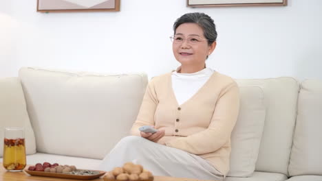 happy middle aged aunt and grandma woman holding remote control, watching tv movie on comfortable sofa at home and pressing the remote control to change channels