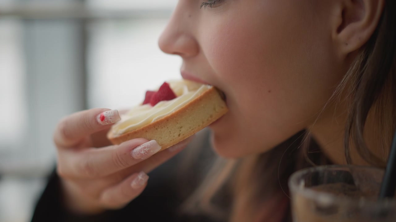 Side-profile close-up of young woman bringing cream and strawberry tart toward lips, taking gentle bite while hand with manicured nails holds pastry near face in cozy cafe with soft indoor light and