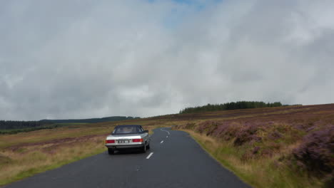 Tiefflug-über-Der-Straße,-Die-Durch-Große-Moorlandschaften-Führt.-Folgenden-Silbernen-Oldtimer-Sportwagen-An-Bewölkten-Tag.-Irland