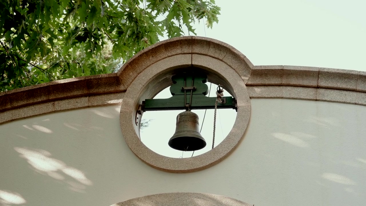 Campana appesa incorniciata da un'apertura circolare in pietra in un muro bianco di chiesa contro il cielo.