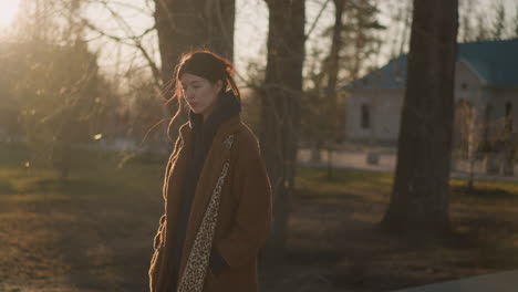 a girl wearing a brown coat, hoodie, and jeans walks through a quiet park during sunset, looking heartbroken. the warm, golden light creates long shadows, adding to the melancholic atmosphere