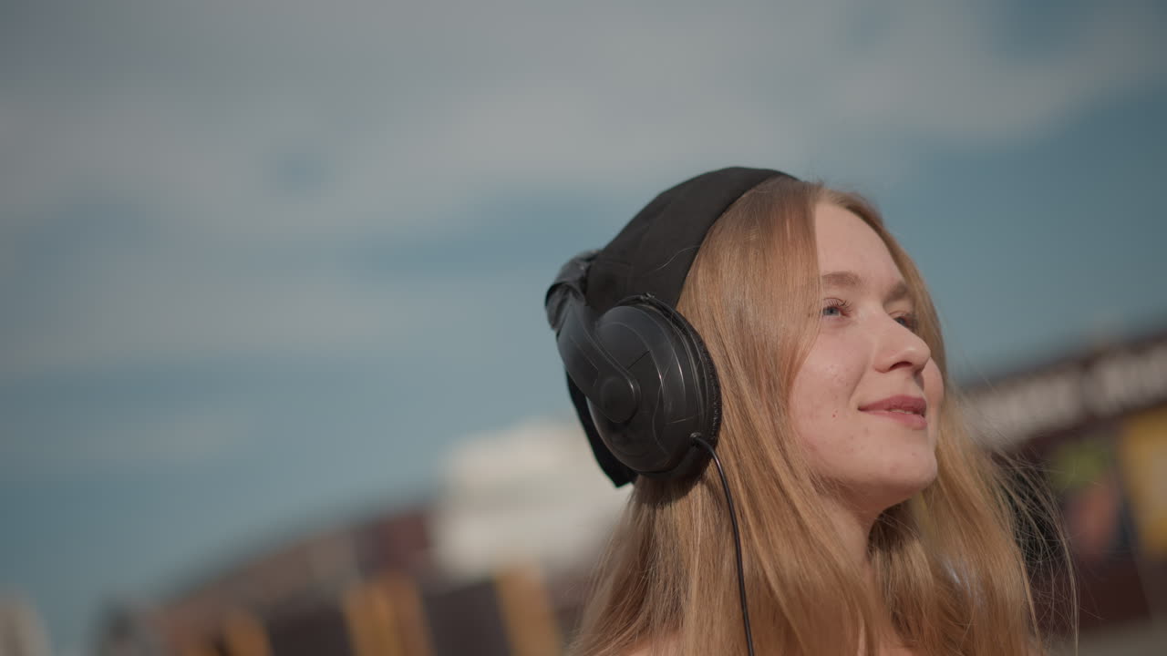 Giovane donna con le cuffie che chiude gli occhi all'aperto, ambientazione urbana, atmosfera serena e sognante, leggero movimento della testa, ascolto riflessivo di una melodia ambientale, luce solare soffusa e nostalgia catturata