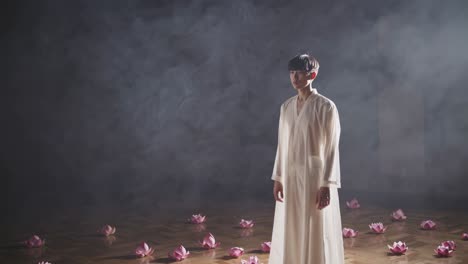young man wearing a white robe is standing in a smoky room with lotus flowers scattered on the floor, creating a serene and meditative atmosphere