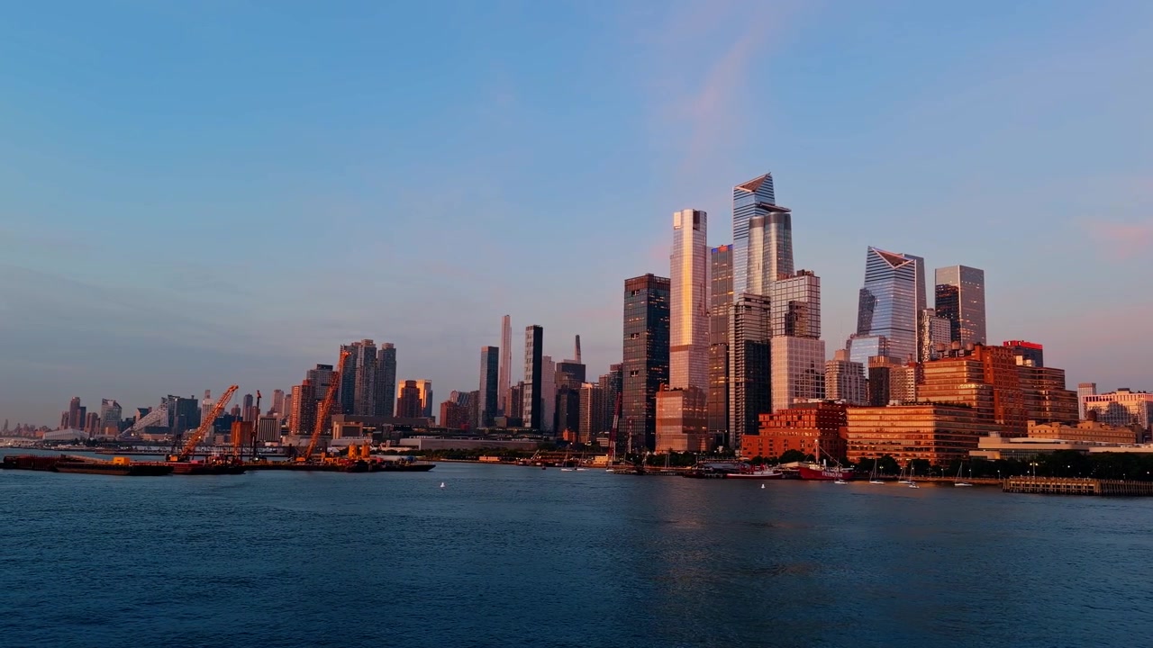 Hudson Yards skyline across Hudson River at sunset, glass towers, harbor cranes and pink clouds.