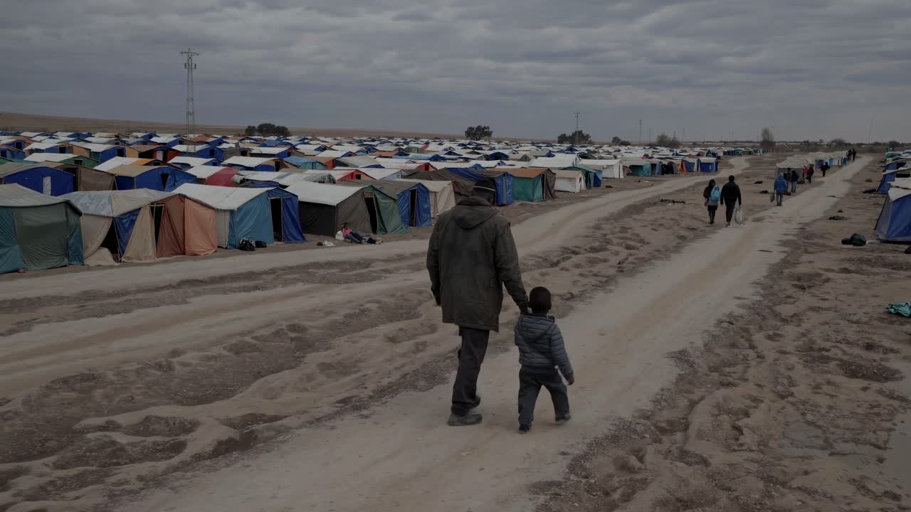 A vast refugee camp with numerous tents. A man and child walk along a dirt path. Overcast sky