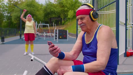 Senior-old-sportsman-grandfather-with-headphones-listening-music-from-mobile-phone-on-playground