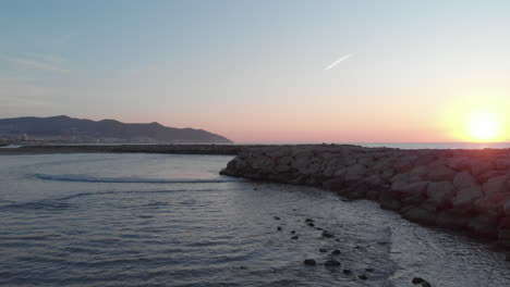 Drone-captures-stone-pear-stretching-into-ocean-as-sun-rises-above-horizon-over-calm-incoming-waves