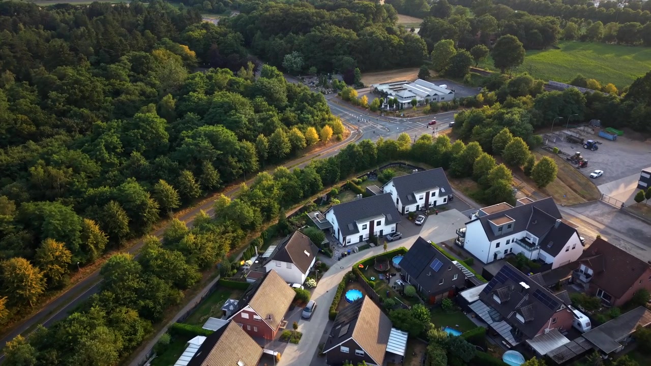 Moderne enfamiliehuse ligger langs en buet blindgyde i en tysk forstad; sorttaget hvide huse med baghavepools grænser op til tæt skov under sommeraftenlys. Luftfoto fra drone.