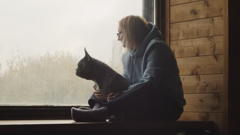 Rothaarige-Frau-Sitzt-Mit-Ihrem-Hund-Auf-Einem-Fensterbrett-Und-Beobachtet-Die-Landschaft