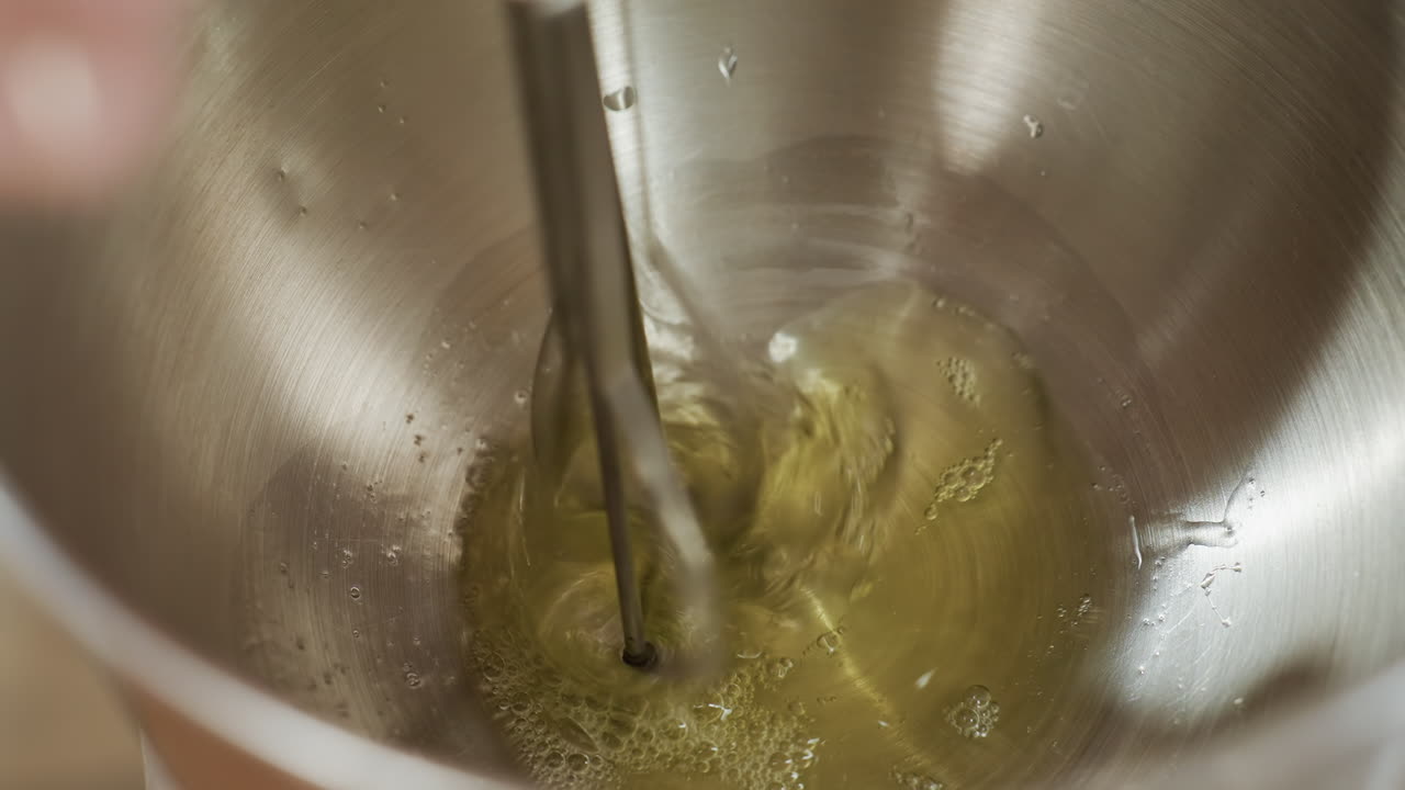 Close up of electric hand mixer blending egg fluid in stainless steel bowl, capturing spinning motion of beaters and liquid swirl as mixing process creates bubbles and smooth consistency