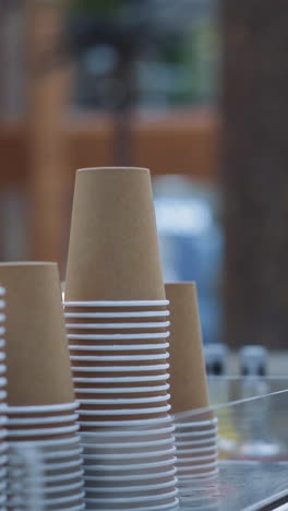 stacks of beige paper cups on display of cafe against blurred view of fountain in spacious hallway of contemporary shopping mall closeup