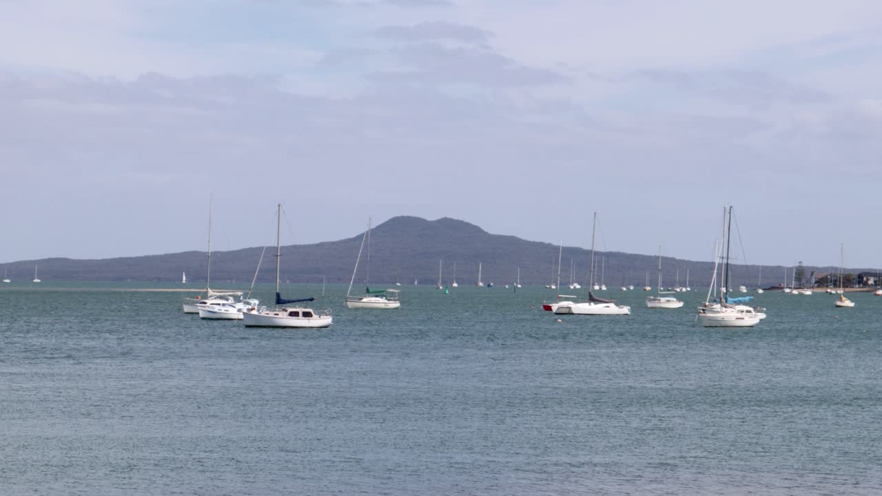 una toma de mano de algunos barcos flotando en un lago con agua azul y un volcán en el fondo en un día ventoso y nublado en auckland, nueva zelanda