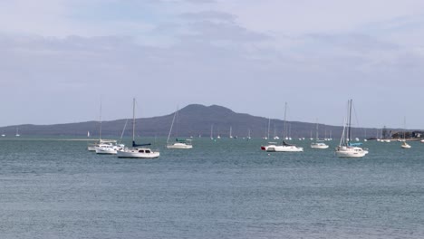 A-handheld-shot-of-some-boats-floating-on-a-lake-with-blue-water-and-a-volcano-in-the-background-on-a-windy-and-cloudy-day-in-Auckland,-New-Zealand