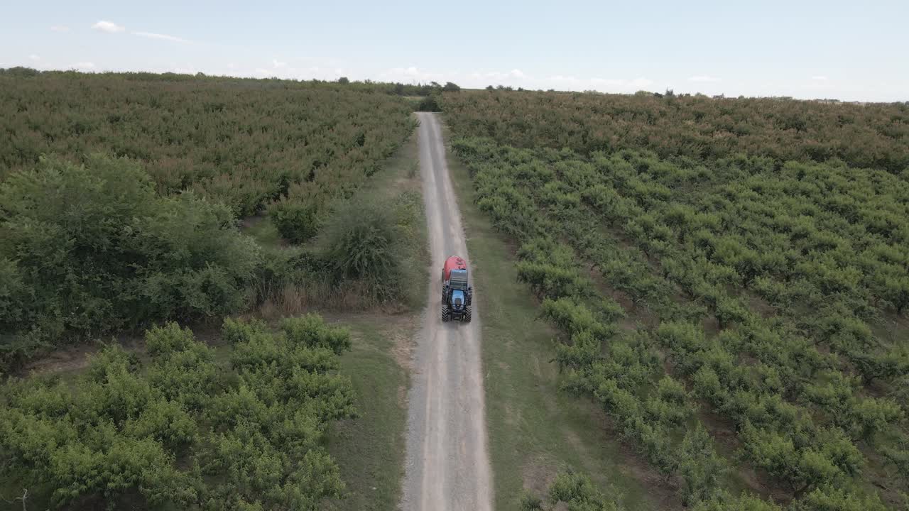 Aerial 4K footage at 50fps of a blue tractor pulling a red sprayer tank along a rural dirt road in a lush orchard, showcasing modern agricultural machinery in a scenic hilly landscape.